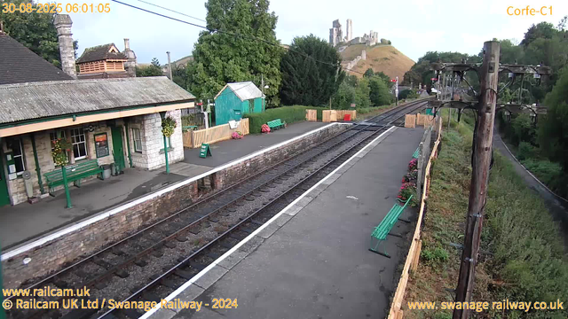 A small train station platform with a building on the left, decorated with hanging flower baskets and green benches. There are flower pots and wooden fences on the platform. Electrical poles with wires run along the right side, and the railway tracks extend into the distance. In the background, there are trees, a small green shed, and a hill with the ruins of a castle at the top. The sky is partly cloudy, suggesting early morning.