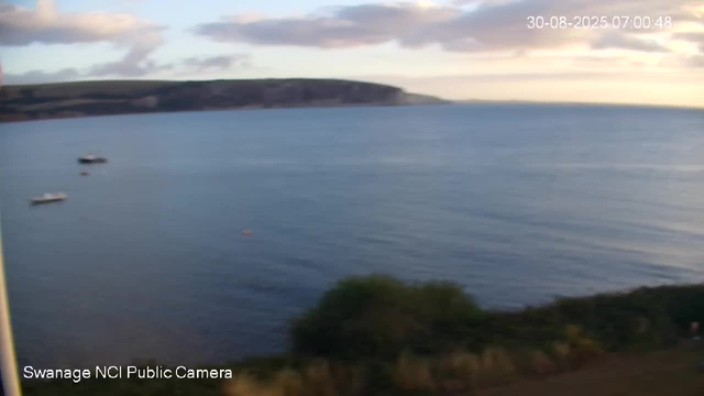 A wide view of a calm body of water during dawn or dusk, with a few small boats floating near the shoreline. In the background, there are low hills or cliffs and a sky with scattered clouds. The timestamp in the top right corner indicates the date and time as August 30, 2025, at 7:00 AM. The bottom left corner is labeled "Swanage NCI Public Camera."