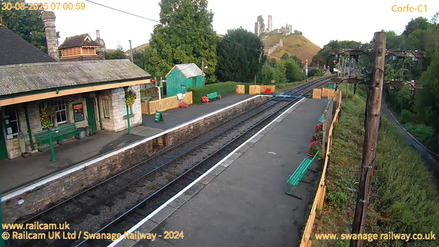 A small train station platform with a green bench, a green shelter with a yellow roof, and decorative flower arrangements in pots. There are yellow barriers on the platform edges and a green sign that says "Way Out." Overhead, electrical wires run from a wooden utility pole. In the background, a hill with ruins or ancient structures is visible, surrounded by trees and greenery. The image has a timestamp of July 30, 2025, at 07:00:59.