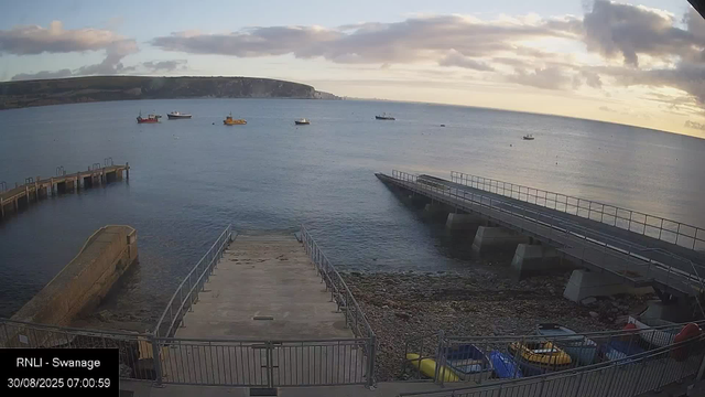 A scenic view of a harbor at sunrise or sunset with calm water, several small boats anchored, and a distant coastline with cliffs. There are two piers extending into the water, one on the left and one on the right, with boats parked along the shoreline. The sky is partly cloudy with a soft, warm light. In the foreground, there is a fenced area with a few kayaks and small boats on the rocky shore. The timestamp indicates it is early morning on August 30, 2025.