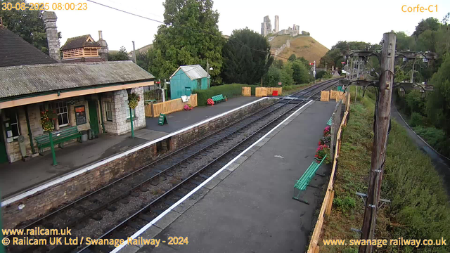 A small, quiet railway station with a single platform and railway tracks running through it. There are green benches, colorful flower arrangements, and a green wooden shed on the platform. A stone building with a tiled roof and hanging flower baskets is adjacent to the platform. In the background, there are sweeping hills with the remains of an ancient castle ruin atop one of the hills. To the right, there is a tall wooden telegraph pole and a foliage-lined road running parallel to the railway tracks. The image appears to be taken early in the morning, as indicated by the timestamp.