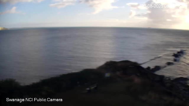 A view of the ocean, with calm water stretching to the horizon under a partly cloudy sky. The image is taken from a high vantage point, showing a rocky shoreline or cliff in the foreground. The sun appears to be setting, casting a warm glow on the right side of the sky. The scene is slightly blurry.