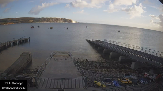 A view of a waterfront scene showing a calm sea with several boats anchored in the distance. There are two piers or docks extending into the water, one on the left and another on the right, with the right pier slightly raised above the water level. In the foreground, there is a small fenced area with boats and possibly kayaks stored inside. A rocky shoreline is visible beneath the piers. In the background, there are cliffs with white chalk formations along the coast, and the sky is partly cloudy with patches of blue visible. The image is timestamped "30/08/2025 08:00:53" and labeled "RNLI - Swanage."