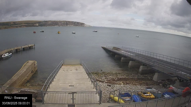 A view of a harbor with several boats anchored in calm water under a cloudy sky. There are two piers extending into the water, one on the left with a small boat docked, and another on the right with a metal railing. In the foreground, there is a concrete slipway and a pebble-strewn shoreline with small boats and colorful kayaks stored along the fence. Distant hills are visible across the water in the background.