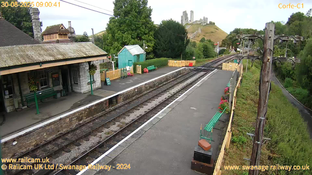 A small train station platform with a stone building on the left, decorated with hanging flower baskets. There are green benches with flower arrangements nearby and a sign that says "WASH OUT." The train tracks run through the center, bordered by a low stone wall. To the right, there are green benches with flowers, a telephone box, and an electric pole with wires. A path runs alongside the station with green trees and hills covered in grass and trees in the background. A castle is visible on a hill in the distant background. The sky is mostly cloudy. The timestamp indicates the photo was taken on August 30, 2025, at 09:00:41.