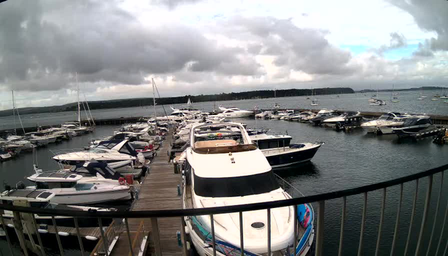 A marina filled with various boats and yachts docked along a wooden pier under a cloudy sky, with more boats sailing in the open water beyond.