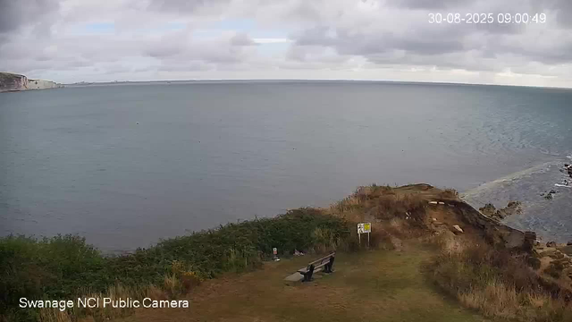A view of a coastal scene showing a calm body of water with a cliff on the left side and cloudy sky above. In the foreground, there is a small grassy area with a bench, some bushes, and a rocky shoreline on the right. The image is timestamped "30-08-2025 09:00:49" and is labeled "Swanage NCI Public Camera."