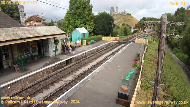 A small train station with a platform beside railway tracks. There are a few people gathered near the station building, which has a stone exterior and a tiled roof. A green bench, a small turquoise shed, and some flower arrangements are visible near the platform. An old wooden telegraph pole stands to the right, with wires extending across the scene. In the background, there is a castle on a hill, surrounded by lush green trees and a partly cloudy sky. The timestamp reads "30-08-2025 10:00:23," and signage indicates it is from Swanage Railway.