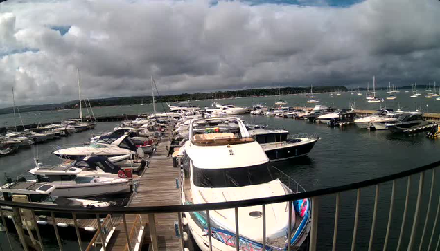 A marina filled with numerous boats and yachts docked along wooden piers under a cloudy sky, with a distant shoreline and a calm body of water in the background.