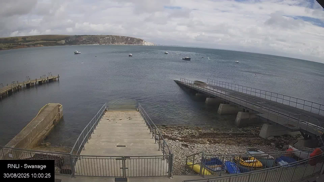 A view of the seaside from a webcam shows a cloudy sky above calm water with several boats floating. In the foreground, there are concrete and metal piers extending into the water, along with a rocky shoreline with colorful small boats. A metal fence surrounds the area, and the distant horizon features white cliffs against the sky. In the bottom left corner, there is a label indicating "RNLI - Swannage" with the date and time "30/08/2025 10:00:25".