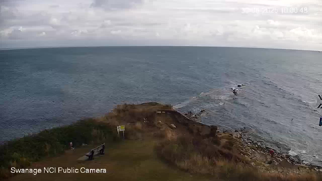A view of a rocky coastline with the ocean in the background. The sky is cloudy, and there are a few benches and signs on a grassy area near the cliff's edge. Waves are gently breaking against the rocks along the shore.