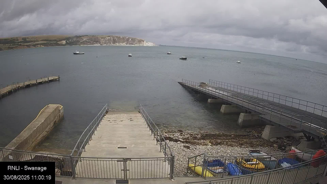 A view of a small harbor with a concrete pier extending into calm water. To the right, there is a sloped boat ramp with a metal railing, leading down to the water. Several small boats are floating in the distance, and the sky is overcast with dark clouds. In the foreground, there is a fenced area with colorful kayaks and small boats. In the background, there are white cliffs and rolling hills under the cloudy sky.
