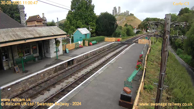 A small railway station platform with a stone building on the left, decorated with hanging flower baskets. There are green benches and flower pots along the platform. To the right, there are electrical poles with wires extending above the tracks. In the background, a hilly landscape with a castle ruin perched on top of a hill can be seen, along with trees, a small green shed, and a wooden fence. The sky is cloudy, and the photo appears to have been taken during daytime.
