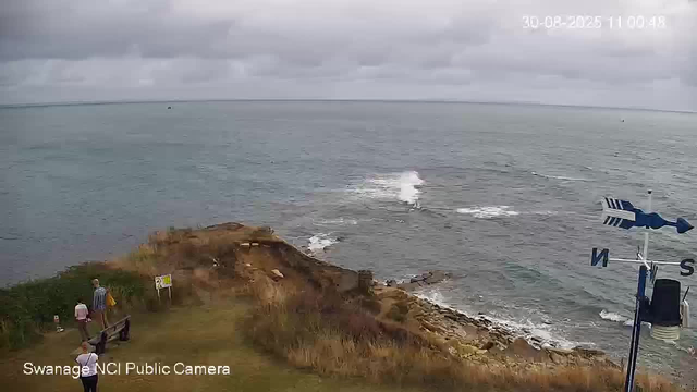 A view of a coastal landscape on a cloudy day. The scene includes a grassy area with a few people standing and sitting on benches near the edge of a cliff. Below is the rocky shoreline with waves gently hitting the rocks. The ocean extends to the horizon under a gray, overcast sky. A weather vane and wind direction indicator are visible on the right side of the image. The timestamp in the upper right corner shows "20-08-2025 11:00:48," and the bottom left has the label "Swanage NCI Public Camera."