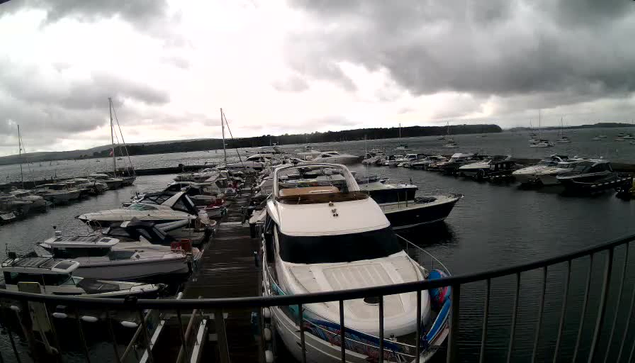 A view of a marina with numerous boats docked on both sides of a wooden pier. The sky is overcast with dark clouds, and a distant shoreline is visible across the water. The railing of the deck from which the photo is taken curves along the foreground.