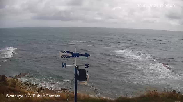 A wide view of the ocean on a cloudy day, with an overcast sky and choppy water. In the foreground, there is a weather vane with a blue arrow and compass directions (N, E, S, W). The shoreline with rocks and some vegetation is visible at the bottom of the image. The timestamp in the top right corner shows "30-07-2023 12:01:40", and the bottom left corner indicates "Swanage NCI Public Camera."