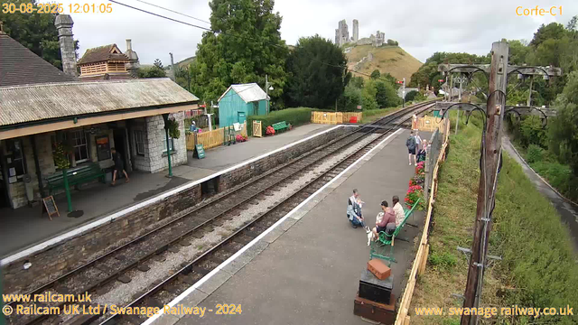 A small train station platform with a stone building on the left, picnic benches, and a turquoise shed in the background. Several people are seated on green benches, some with luggage or bags. A few people are walking along the platform, and a couple of individuals are engaged in conversation. The railway tracks run through the middle of the image, bordered by a yellow safety fence. On the right side, a wooden fence separates the platform from a path lined with greenery and a series of wooden utility poles supporting electrical wires. In the distance, a castle sits atop a hill, partially obscured by trees, under an overcast sky. The timestamp on the image shows 12:01:05 on August 30, 2025.