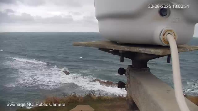 A view of the ocean with waves crashing against the shoreline. In the foreground, there is a mounted device or sensor attached to a wooden platform. The sky is overcast with gray clouds, and there is a distant landmass or headland on the horizon. The image appears to be taken from a webcam situated near the coast. The timestamp shows it was captured on August 30, 2025, at 1:00 PM.