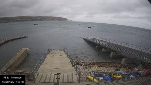 A view of a harbor with several small boats floating on calm water under a cloudy sky. In the foreground, there are concrete platforms and metal railings, with colorful kayaks and small boats stored along the edge. A rocky shoreline is visible at the bottom and a distant landmass with cliffs can be seen across the water.
