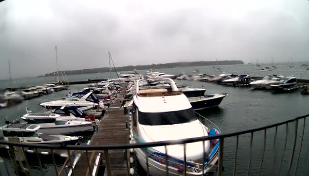 A marina with numerous boats docked along a wooden pier. The boats vary in size and style, and are moored on calm water under an overcast sky. In the background, there is a distant shoreline with some land or islands.