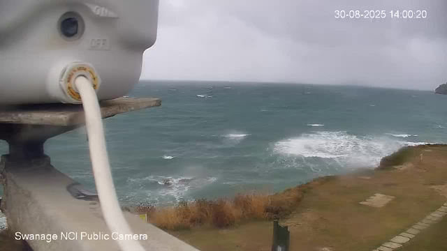 A view of the ocean and coastline from a webcam on a cloudy day. In the foreground, part of a white surveillance camera with a visible cable and mounting base is seen. The sea has whitecaps, indicating windy conditions, and the coastline appears rocky with some vegetation. The image is timestamped "30-08-2025 14:00:20" and marked "Swanage NCI Public Camera" at the bottom.