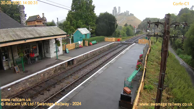 A small train station with a platform next to railway tracks. There are two people waiting under the shelter, which has a hanging flower basket and a blackboard sign. The platform has benches, flower pots, and a small green building in the background. A wooden fence separates the platform from a grassy area with trees. In the distance, a hill with a few buildings on top is visible, with some towers or structures on the summit. An electric pole with wires runs along the right side of the platform. The image appears to be taken on a rainy day, as the platform surface looks wet. The timestamp shows August 30, 2025, at 14:00.