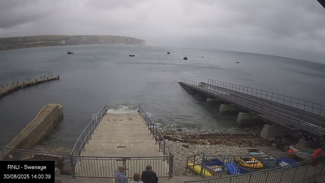 A panoramic view of a cloudy day at a coastal area with multiple boats floating on calm water. In the foreground, a small pier extends into the sea with a gentle wave lapping against it. To the right, a ramp leads down to the water, supported by concrete bases. Below, there are paddle boats and small watercraft parked behind a metal fence, with a few people standing near the fence observing the scene. In the distance, a cliff with some greenery is visible over the horizon. The image has a timestamp at the bottom left indicating it was captured on August 30, 2025, at 14:00:39.