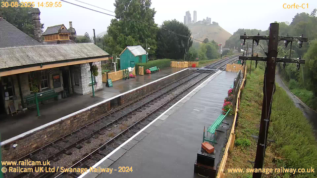 A small train station with a wet platform on a rainy day. There are green benches along the platform, some flower pots with pink flowers, and a small green shed. An old stone building with a sloped roof and a chimney is on the left. Tall trees surround the station, with rolling hills and a castle or large building on a hill in the background. Overhead power lines run above the train tracks, which extend into the distance. The sky is cloudy and misty.