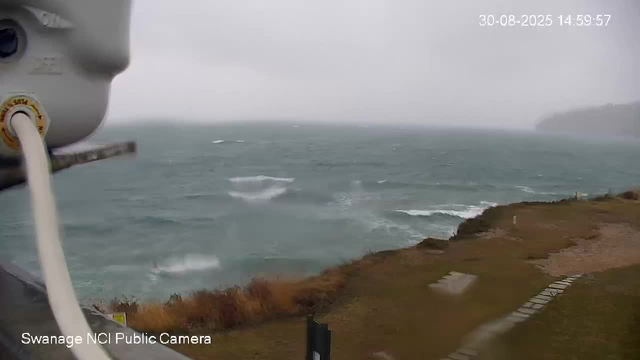A view from a webcam showing a stormy sea with choppy waves crashing against a rugged coastline. The sky is overcast and gloomy, with rain continuing to fall. In the foreground, part of the weather station or camera equipment is visible on the left side, and water droplets are seen on the camera lens. A timestamp in the top right corner indicates the date and time as August 30, 2025, at 14:59:57. A small caption at the bottom left reads "Swanage NCI Public Camera."
