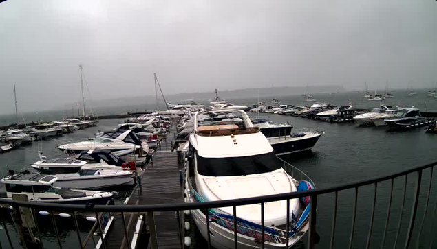 A marina with multiple boats and yachts docked along wooden piers under an overcast sky. The water appears calm, and the scene is viewed from behind a curved metal railing. Some boats are large and modern, while others are smaller. The horizon shows more boats faintly visible through the foggy weather.