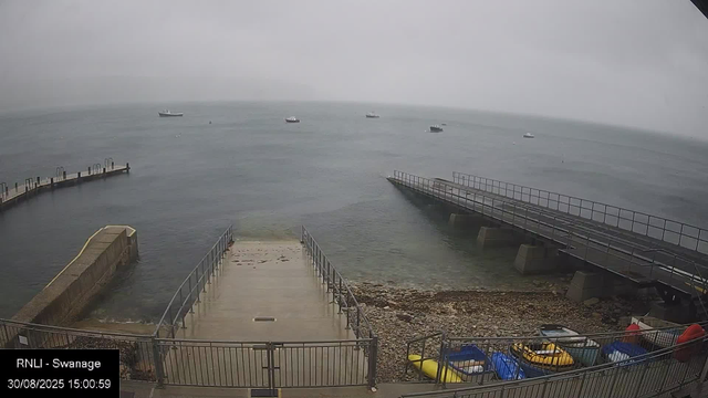 A view of a harbor on a cloudy day with several boats floating in the water. There are two concrete piers extending into the sea, one in the foreground and another on the right. At the bottom of the image, there is a small section with colorful kayaks and boats stored behind a metal fence. The timestamp on the image indicates it was taken on August 30, 2025, at 15:00:59.