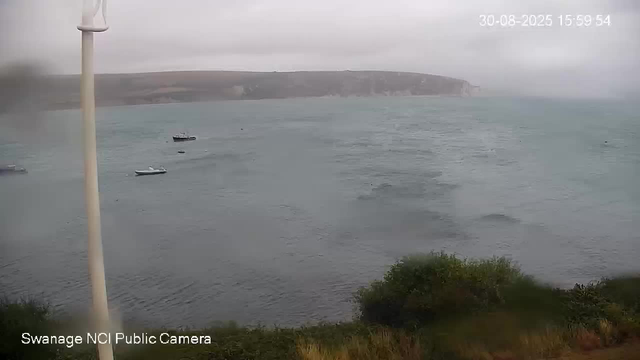 A view of a cloudy sky over a body of water with several small boats floating. In the distance, there are cliffs or hills along the shoreline. In the foreground, there is some greenery and bushes. There is also a vertical pole on the left side of the image. The timestamp in the top right corner shows "30-08-2025 15:59:54."