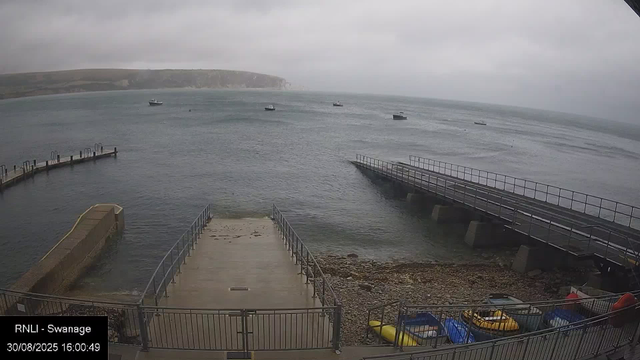 A view of a cloudy, overcast seaside scene with a small dock extending into the water. Several boats are anchored in the distance on the calm sea. In the foreground, there is a concrete slipway leading into the water, with a metal railing along its sides. To the right, a larger ramp extends into the water, supported by concrete pillars. At the bottom right corner, a small collection of colorful kayaks and watercraft are visible on the rocky shore. The background features a distant cliff or headland under a gray sky.