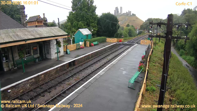 A small railway station platform with two green benches, flowerpots with pink flowers, a small green shed, and a brick building with a sloped roof. The railway tracks run parallel to the platform, and there are tall, dark utility poles with wires overhead. In the background, a hill with a castle ruin on top and some trees surrounding the scene. The sky is overcast.