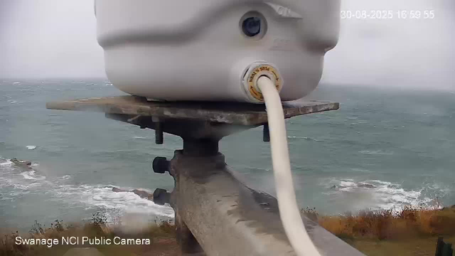 A close-up view from a webcam shows a white device mounted on a metal pole, overlooking a coastline with the ocean and waves crashing onto the shore. The device has a cable connected to it, with an overcast sky in the background. The timestamp on the image is "30-08-2025 16:59:55," and the bottom left corner indicates it is from "Swanage NCI Public Camera."