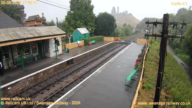 A small railway station platform with a stone building, green benches, and flower planters. The platform is wet, suggesting recent rain. There are overhead electric lines and a posts on the right side with mounted electrical equipment. In the background, a hilly landscape with some tall buildings visible through mist. An orange fence runs along the edge of the platform, and a pathway with trees and greenery is on the far right side. The timestamp indicates it is 5:00 PM on August 30, 2025.