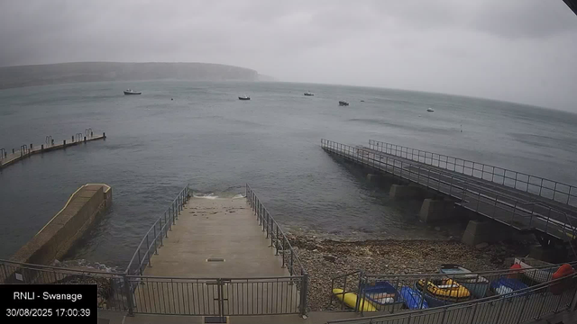 A view of a small harbor on a cloudy day. There are three boats anchored in the water, and two piers extending into the sea on either side. The shoreline has rocks and pebbles, with several kayaks and small boats stored in a fenced area at the bottom of the image. The sky is overcast, creating a grey and calm atmosphere.