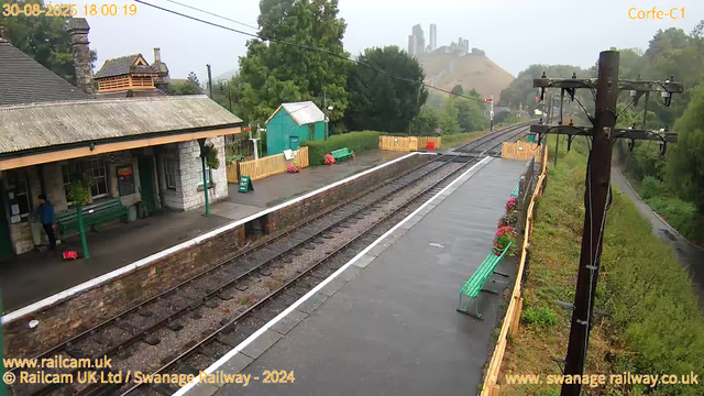 A small train station platform with a stone building on the left, featuring a green bench, potted plants, and a person standing near the building. The platform has two green benches, flower arrangements, and yellow fencing along the edge. Railway tracks run parallel to the platform, with a utility pole and power lines on the right side. In the background, there are lush green trees, a hill with a castle-like structure on top, and a misty sky. The timestamp shows "30-08-2025 18:00:19".