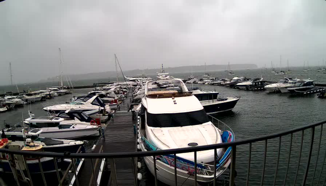 A harbor filled with numerous moored boats and yachts, viewed from a wooden dock structure with black metal railings. The scene is overcast with a cloudy sky and misty atmosphere, and the water appears calm with boats floating gently. In the background, there is a distant shoreline with more boats visible farther out in the water.
