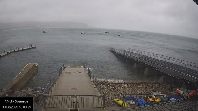 A view of a cloudy, overcast day at a seaside location with a calm sea. Several boats are floating in the water in the distance. There is a small concrete pier with metal railings extending into the water in the foreground, along with a boat ramp and a parking area containing colorful pedal boats and other small inflatable watercraft. The image includes a timestamp and location label reading "RNLI - Swannage" and the date "30/08/2025 18:00:29."