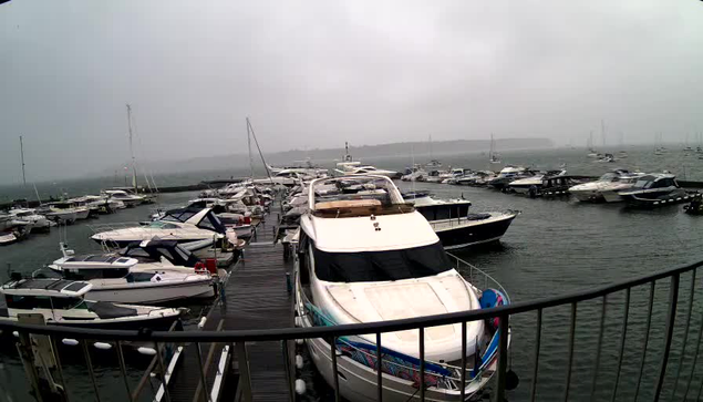 A marina with numerous boats and yachts docked along wooden piers on a cloudy, foggy day. The water appears calm, and the horizon is barely visible through the mist. A curved metal railing is in the foreground, suggesting the photograph was taken from a vantage point on a balcony or walkway overlooking the marina.