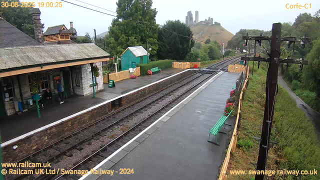 A small train station platform with a building on the left side, where two people are sitting on benches. There are flower planters along the edge of the platform, with purple and pink flowers. The train tracks run down the center of the image, disappearing into the distance. To the right, there is a tall wooden utility pole with wires extending across the image. Beyond the platform, there is a green pathway with trees and bushes on the right, and in the background, a hill with an old castle ruin atop it. The sky is overcast. The timestamp indicates the image was captured on August 30, 2025, at 7:00 PM.
