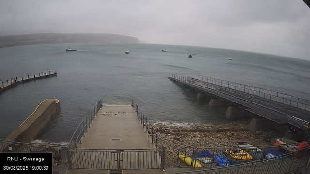A view of a small harbor with cloudy skies. Several boats are floating in the water in the distance, and a pier extends into the sea. In the foreground, there is a concrete platform with metal railings, adjacent to a rocky shoreline. At the bottom of the image, there are colorful kayaks or small boats stacked in a fenced area. The scene appears overcast and slightly foggy.