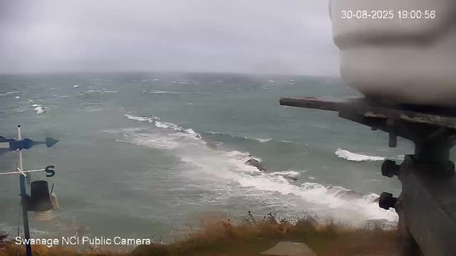 A view of the ocean on a cloudy day, with waves crashing against rocks near the shoreline. A weather vane with a windsock is visible on the left, and some equipment or part of a structure is seen on the right. The timestamp in the top right corner indicates the date and time as August 30, 2025, at 19:00:56. The camera is labeled "Swanage NCI Public Camera" at the bottom left.