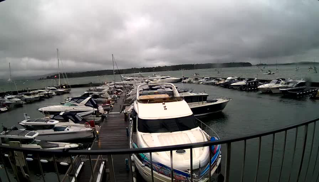 A marina filled with numerous boats and yachts docked along wooden piers under a cloudy, overcast sky. The water appears calm, and there is a distant shoreline visible on the horizon. In the foreground, a large white yacht is prominently positioned, with a railing partially framing the scene.