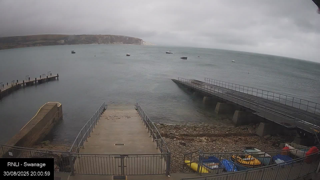 A view of a harbor on a cloudy day, with a concrete pier extending into the water in the foreground. Several boats are anchored in the calm sea, and a rocky shoreline is visible beneath the pier. In the distance, there are cliffs and a low-lying landmass under an overcast sky.