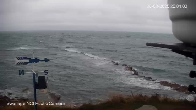 A view of the ocean on a cloudy day, with waves crashing against rocks along the shoreline. There is a weather vane with directional indicators (N, S), and part of the webcam's mount or camera equipment visible on the right side of the image. The timestamp shows "20-08-2025 20:01:03."