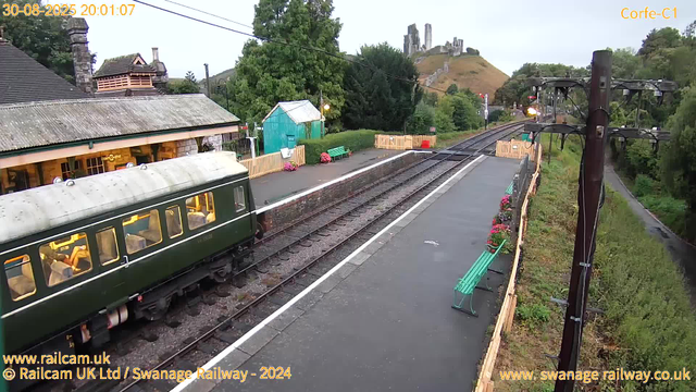 A vintage green train with a rounded roof is stopped at a small railway station platform, illuminated from inside. The platform has green benches and colorful flower pots. A small green building is visible behind the platform. In the background, there is a hill with the remains of a castle or fortress on top, and a large tree is to the left. The sky is overcast, and the scene is captured in the early evening, as indicated by the timestamp. There is also a wooden fence running along the platform edge and electrical poles with wires overhead.