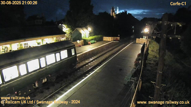 A black train is stopped at a small station platform at night. The station is illuminated by warm yellow lights, with a small green building and a green bench visible on the platform. The surrounding area is dark, with a hill and a faintly lit structure or castle on top of the hill in the background. The railway tracks run parallel to the platform, and there are wooden fences and utility poles along the side. The sky is mostly dark with some clouds illuminated faintly by the moon or nearby lights. The timestamp on the top left indicates 21:00:26 on August 30, 2025.