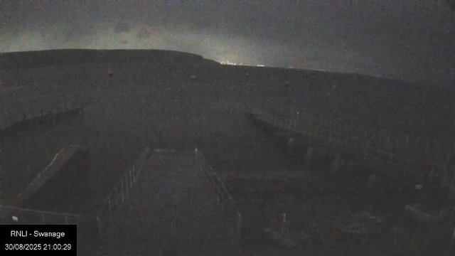 Nighttime view of a bridge spanning across a river, with a dark sky overhead and faint rain visible in the air. The bridge is illuminated by some lights, and the area appears calm with no visible activity on the water. In the lower left corner, there's a timestamp and label indicating the location as Swansea and the date as August 30, 2025, at 21:00:29.